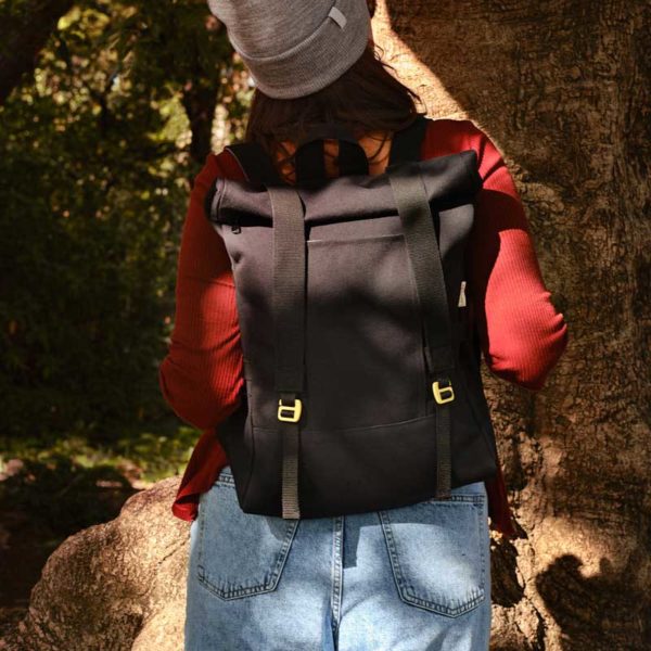 woman standing in front of a tree, she carries black recycled_roll_top_backpack on her shoulders