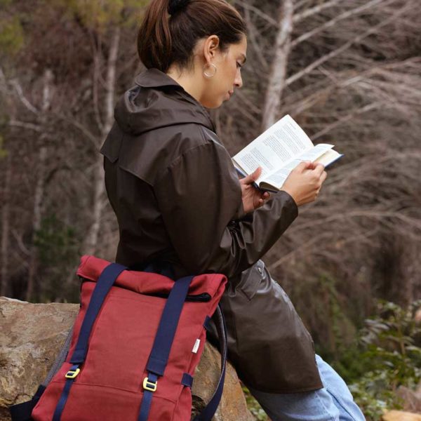 woman reading a book, next to her there is ared recycled roll top backpack